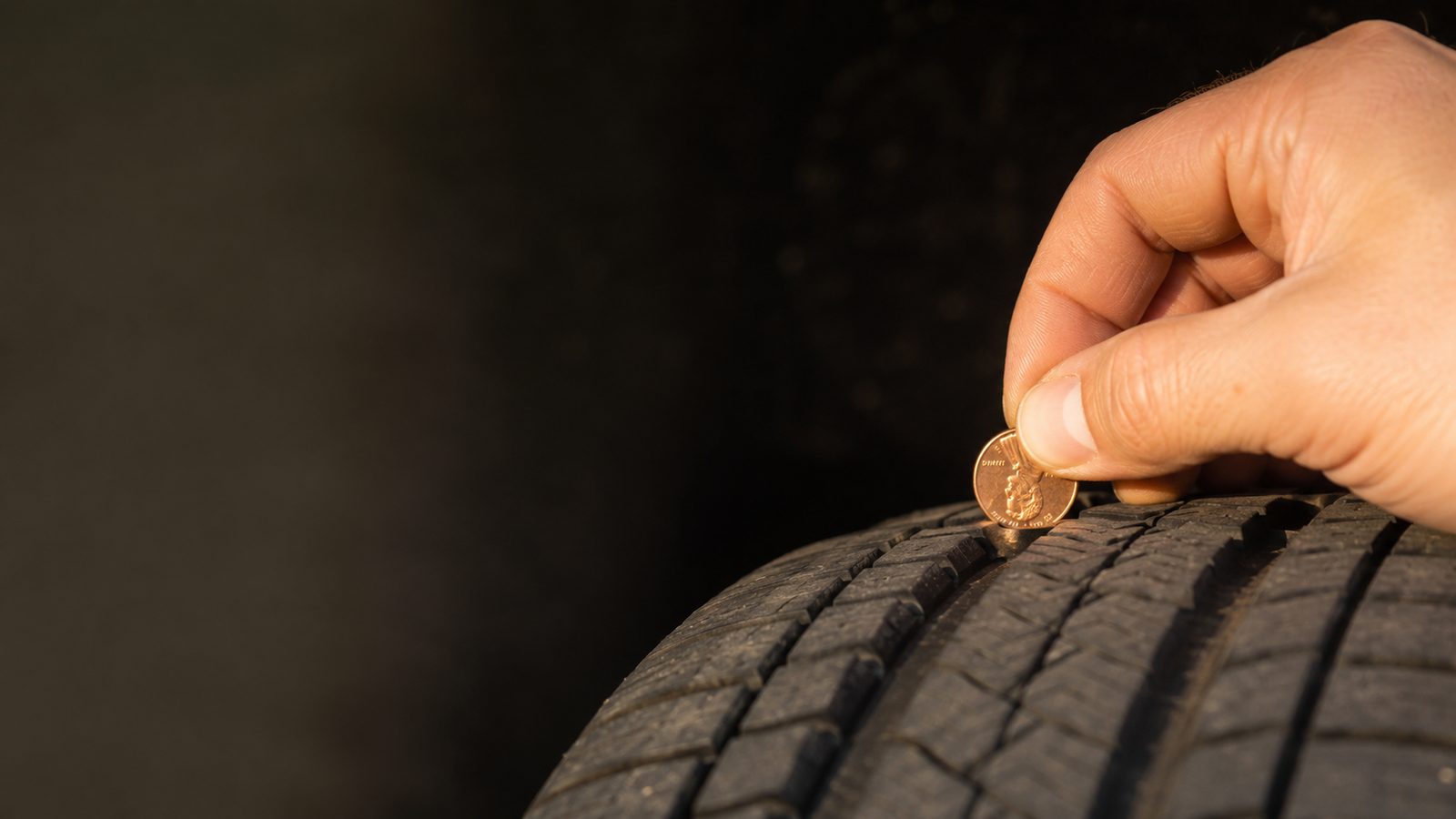 A hand inserting a U.S. penny into a car tire's tread groove with Lincoln's head pointing down, demonstrating the penny tread-depth test